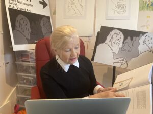 This is a photograph of Artist Clare Mc Laughlin taken in her art studio, during a zoom presentation to the Seen - Unseen participants. Clare is in the foreground seated in a red chair talking to her laptop with a copy of her thesis in her left hand. She is reading from this. In the background of the photograph are various artworks and line drawings from Seen - Unseen.